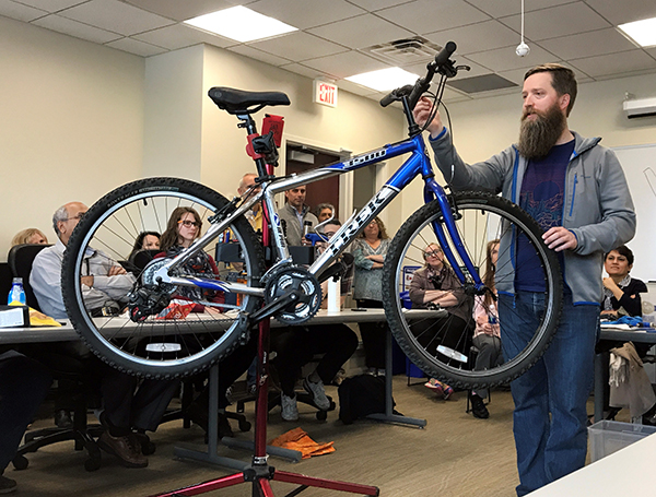 Man next to a bike on a workstand. His right hand is pointing at the shifter pod, or device used to change gears on a bike, as he explains common issues with that particular assembly.